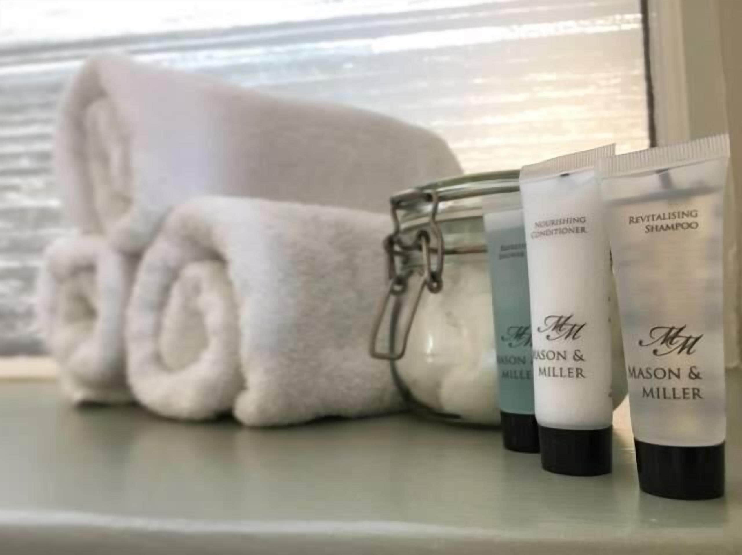 A glass jar with toiletries, including Revitalising Shampoo, next to a rolled white towel on a windowsill.