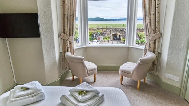 A hotel room with a large window offering a view of a landscaped area and a stone bridge. There are two upholstered chairs and a bed with neatly folded towels. - The Commodore Inn (Grange-over-Sands)