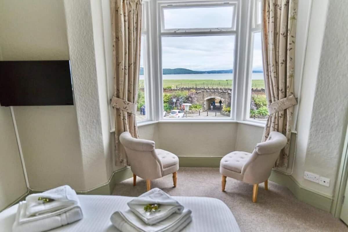 A hotel room with a large window, two chairs, a bed with towels, and a flat-screen TV.