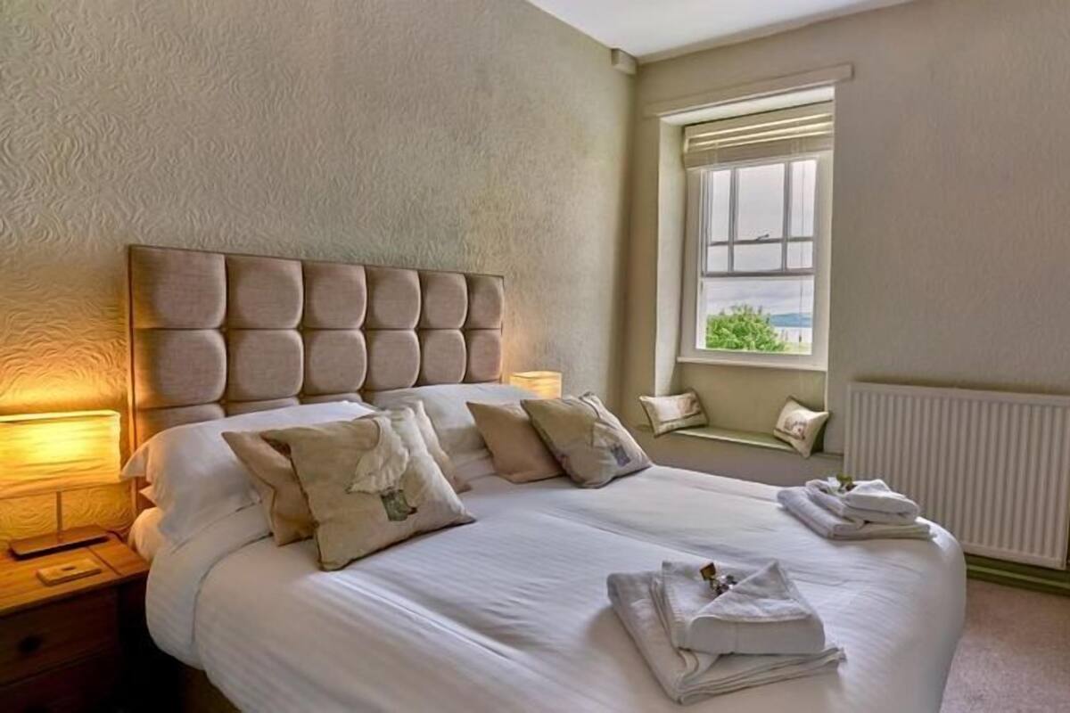 A neatly made bed with pillows and folded towels, a headboard, bedside lamps, a window with a view, and a radiator.