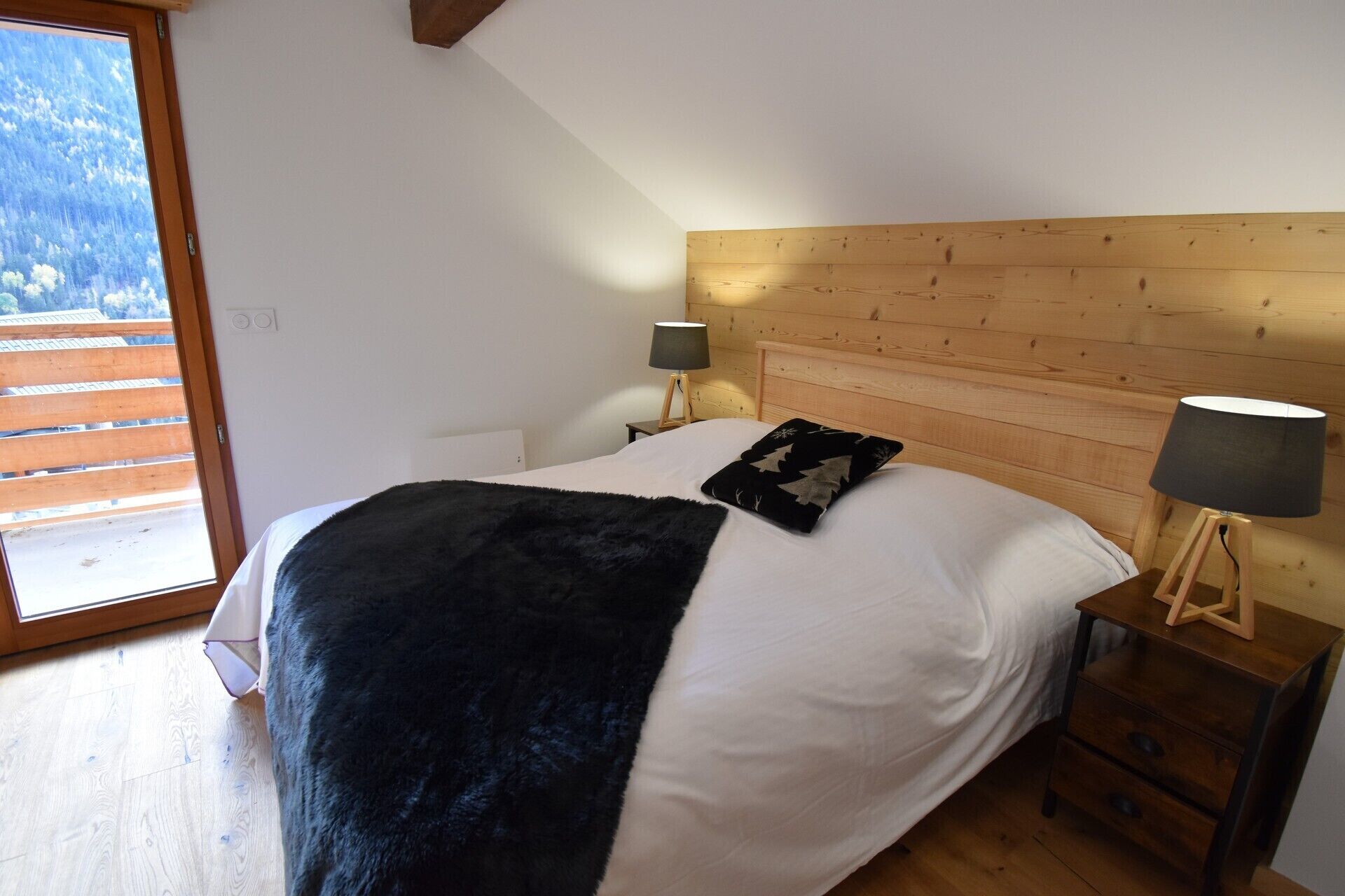 Une chambre à coucher avec une tête de lit en bois, un grand lit recouvert d’une couverture noire et blanche, deux lampes de chevet et une vue sur l’extérieur par une fenêtre.
