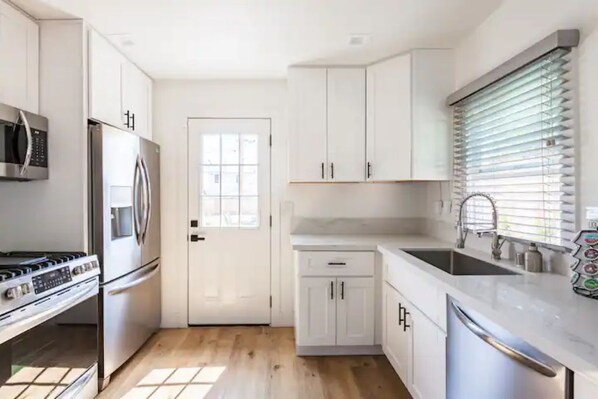 Une cuisine moderne avec des meubles blancs, des appareils en acier inoxydable et une grande fenêtre équipée de stores.