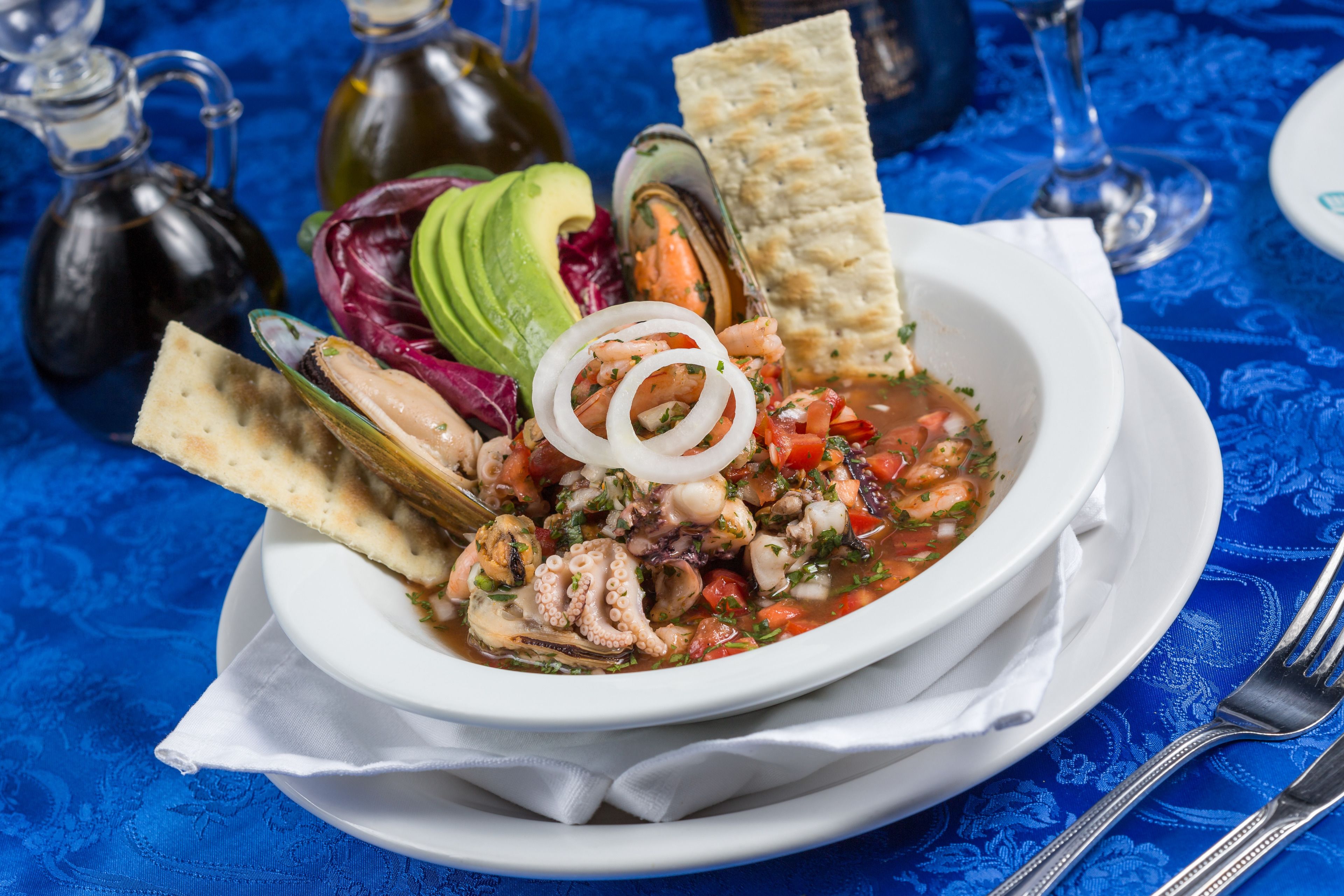 Un tazón de sopa de mariscos con diferentes ingredientes, como aguacate, aros de cebolla y un trozo de pan, sobre un plato blanco.