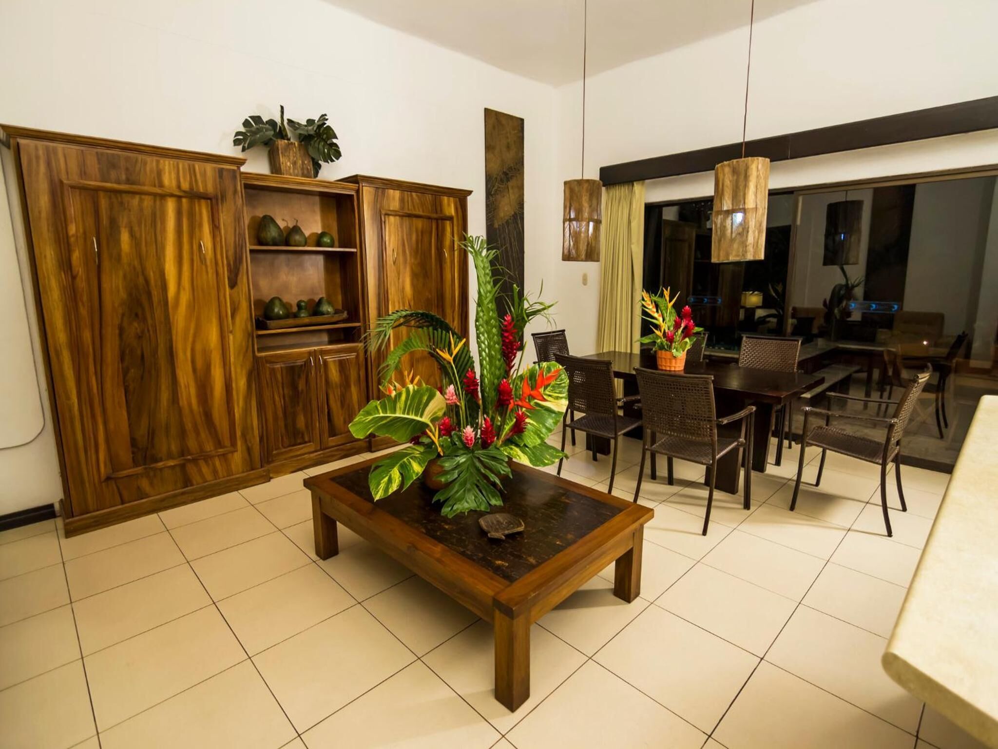 A spacious living area with a wooden coffee table, dining table, and chairs. There is a large wooden cabinet with shelves and a potted plant.