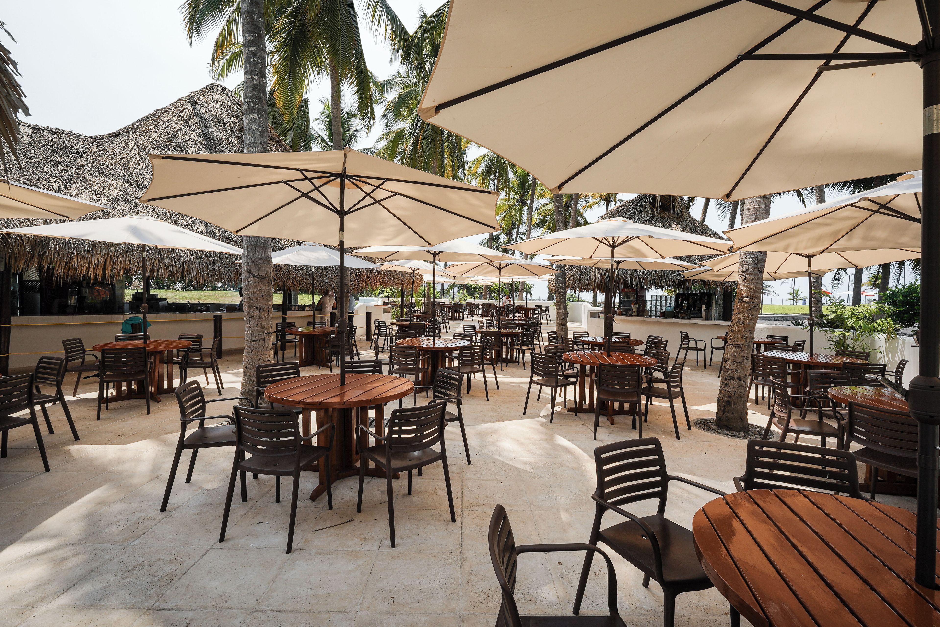 An outdoor café area with multiple tables and chairs, shaded by large umbrellas, and surrounded by palm trees.