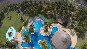 An aerial view of a resort with a central swimming pool surrounded by palm trees and a thatched-roof structure.