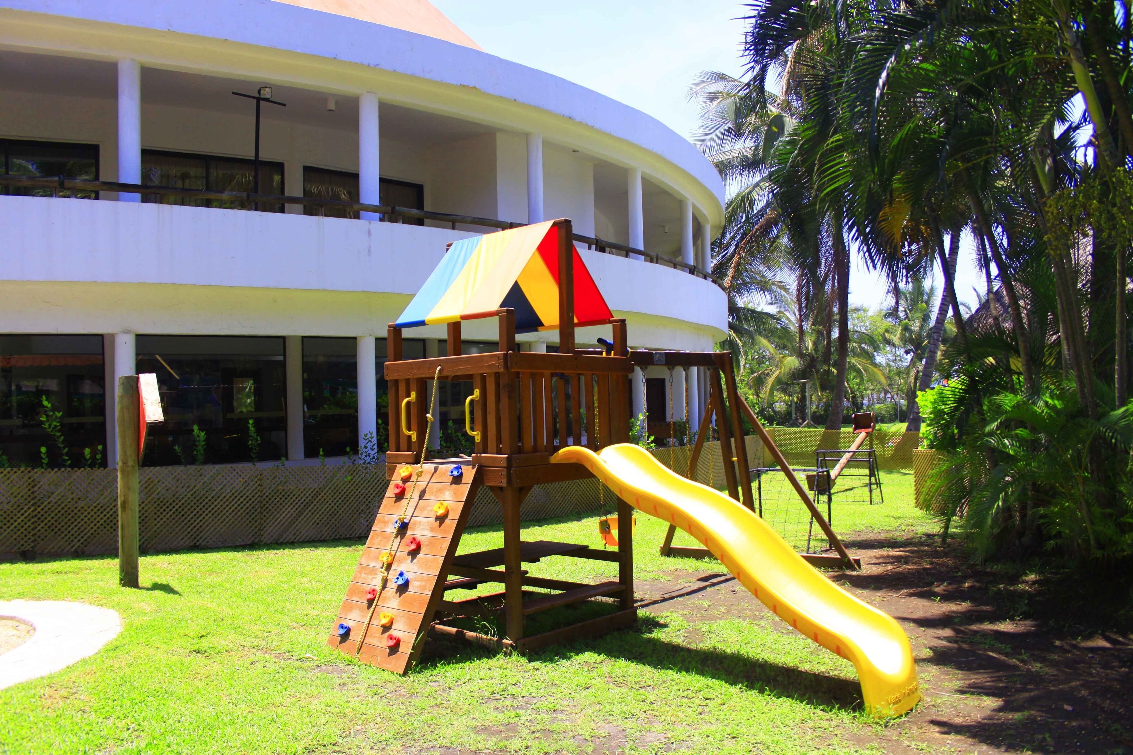A playground with a slide and climbing wall, surrounded by a grassy area and palm trees.