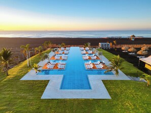 Piscina de resort con varias pistas de natación, rodeada de sillas de descanso y palmeras, con vista a una playa.