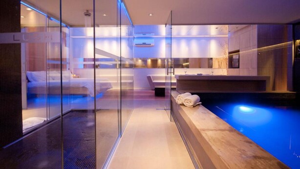 A modern hotel room with a glass partition, a bed, a small seating area, a vanity with a mirror, and a pool area. - Lush Motel Ipiranga (São Paulo)