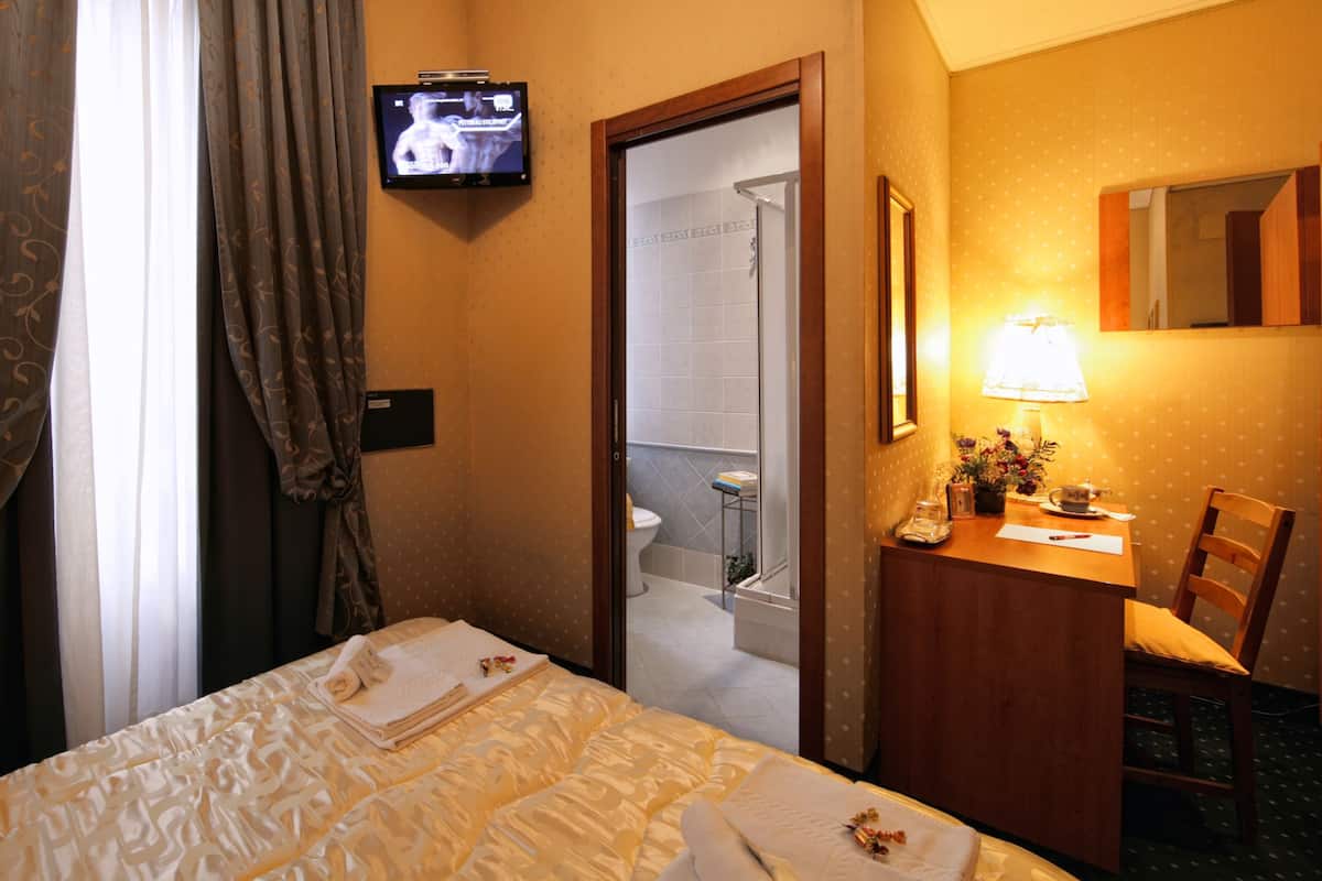 A hotel room with a bed, a desk with a lamp, a television on the wall, and a bathroom visible through an open door.