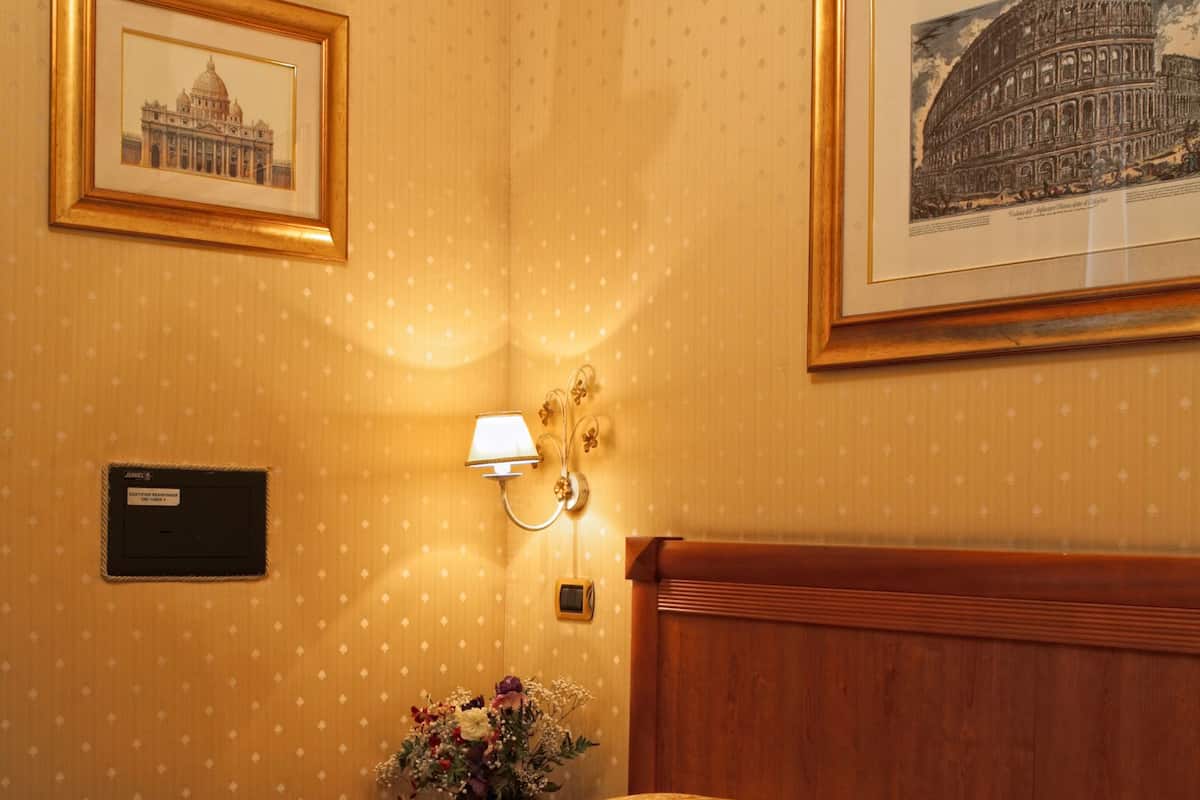 A hotel room with a bed, a nightstand with a lamp, a framed picture of a building, and a nameplate on the wall.