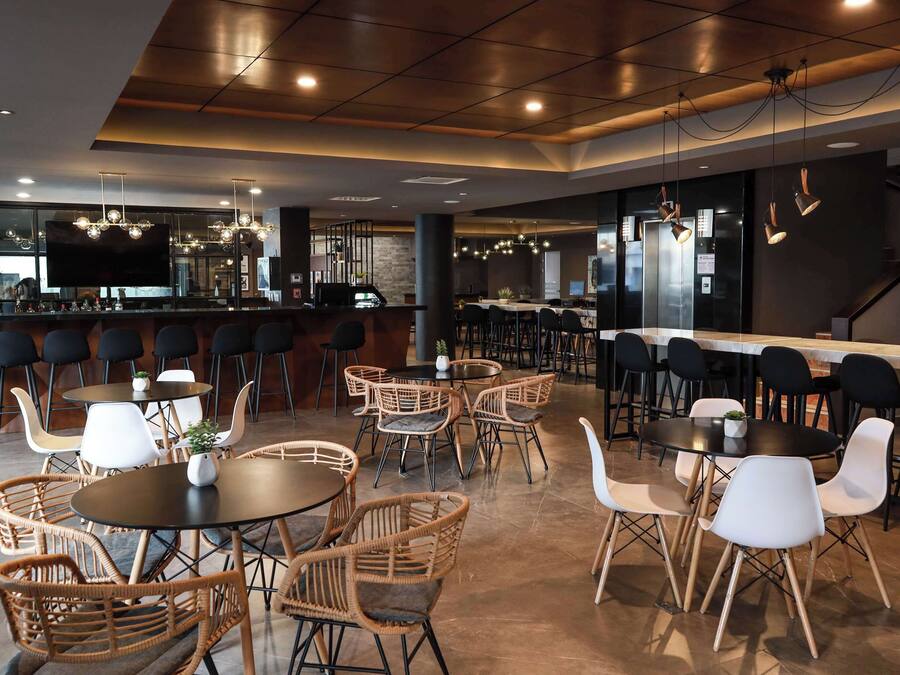 A modern restaurant with round tables and wicker chairs, a bar area with stools, and a visible kitchen in the background.