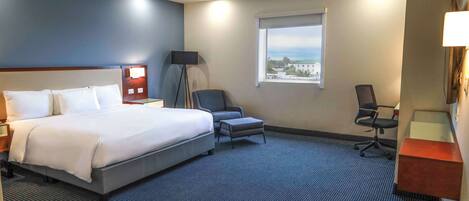 A hotel room with a large bed, a desk, a chair, a small table, a lamp, and a window with a view.