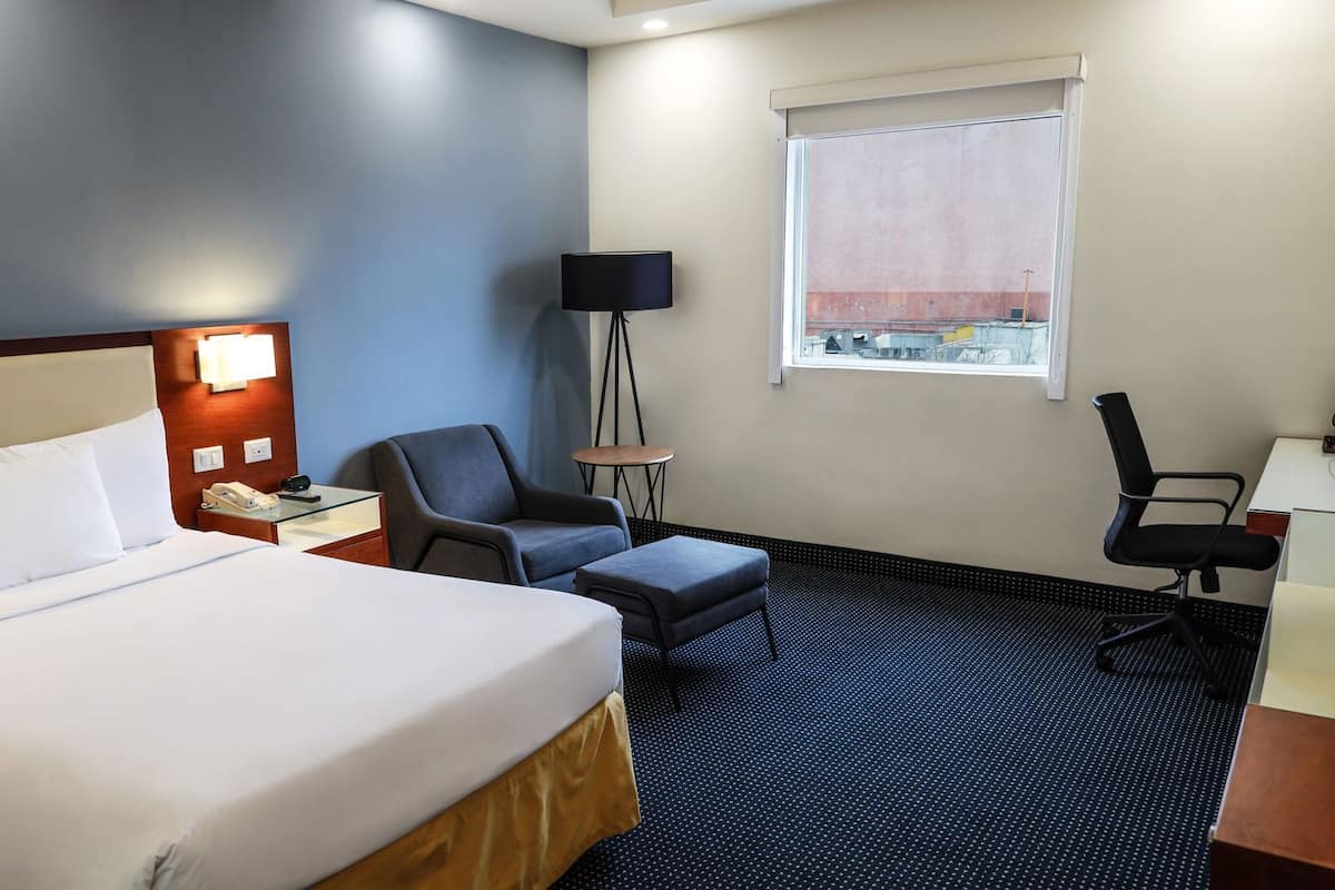 Una habitació d'hotel amb un llit, escriptori, cadira, taula petita i una làmpada.