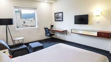 A hotel room with a large bed, a desk with a chair, a TV, a window with a view, and a small table with a lamp.