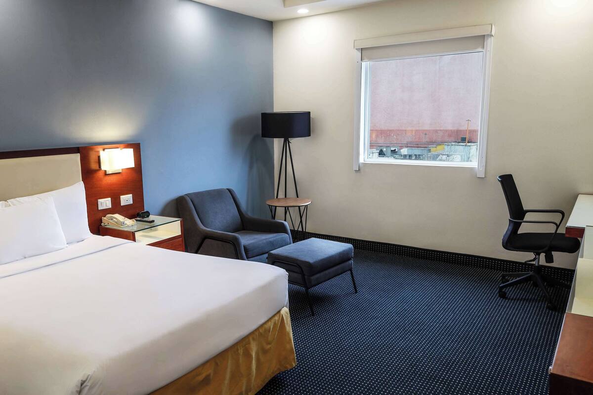 Habitació d'hotel amb llit, escriptori, cadira, taula petita, làmpada i finestra.