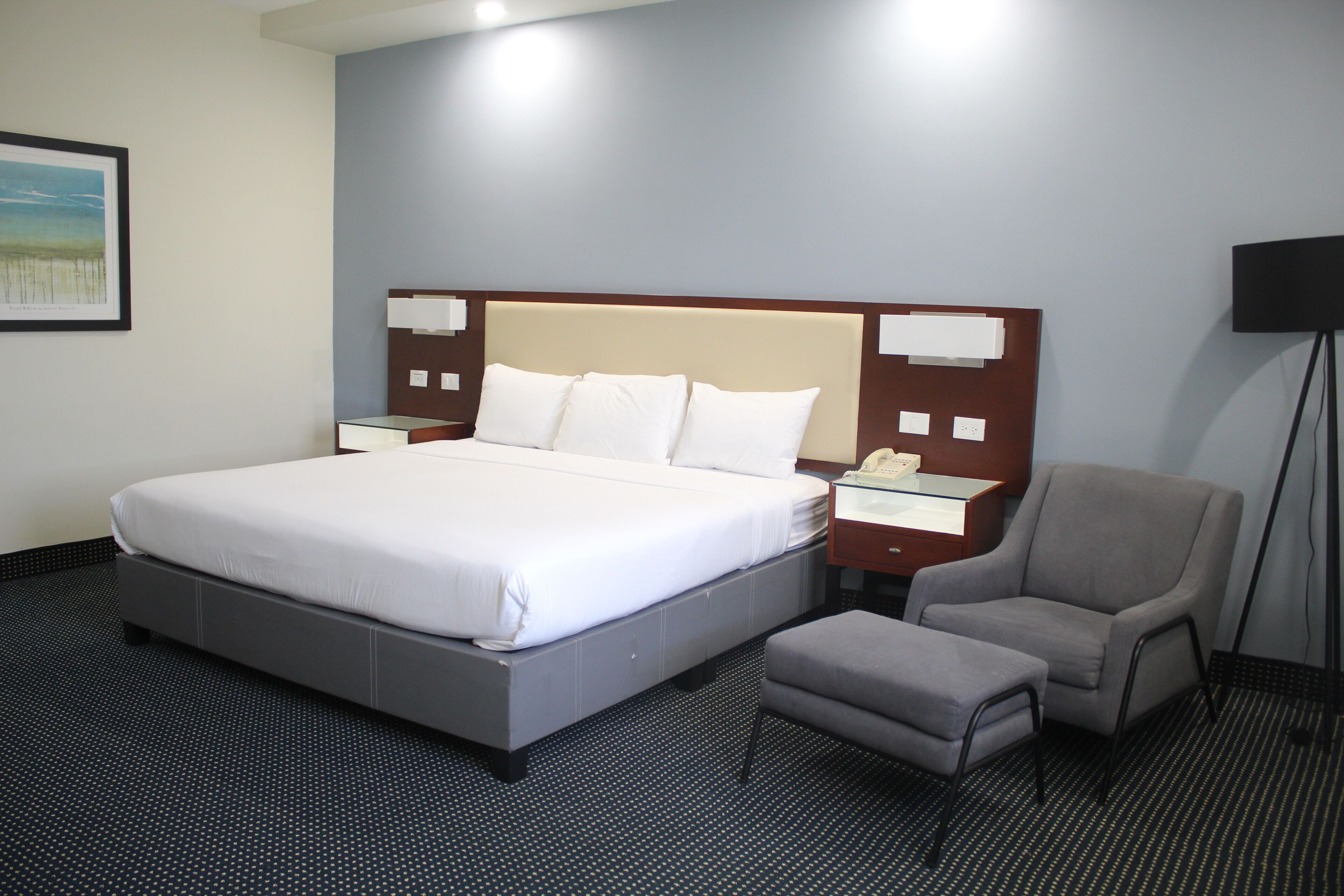 A hotel room with a large bed, a bedside table with a phone, a chair, and an ottoman.