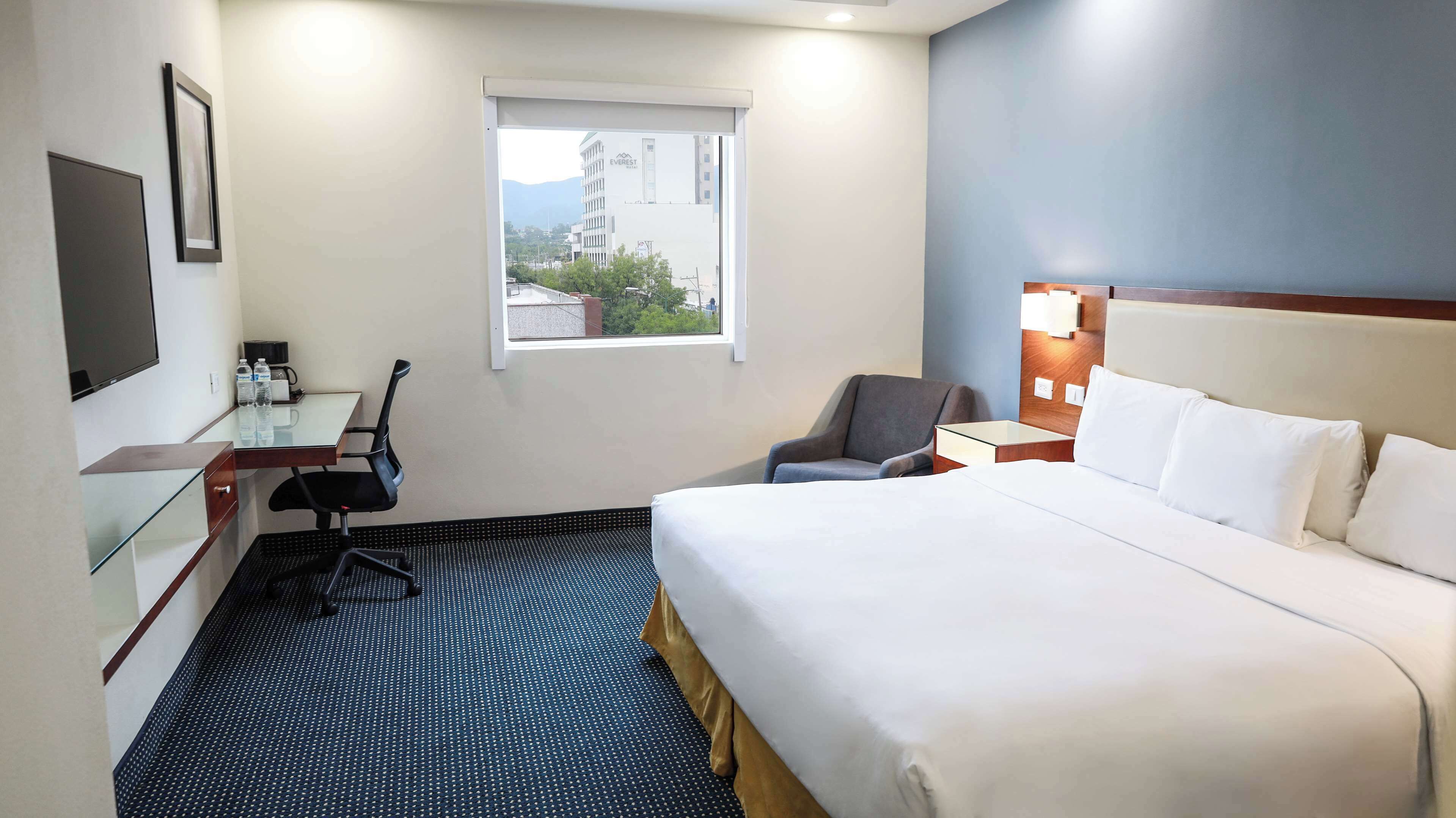 A hotel room with a bed, a desk with a chair, a TV, and a window with a view of buildings.