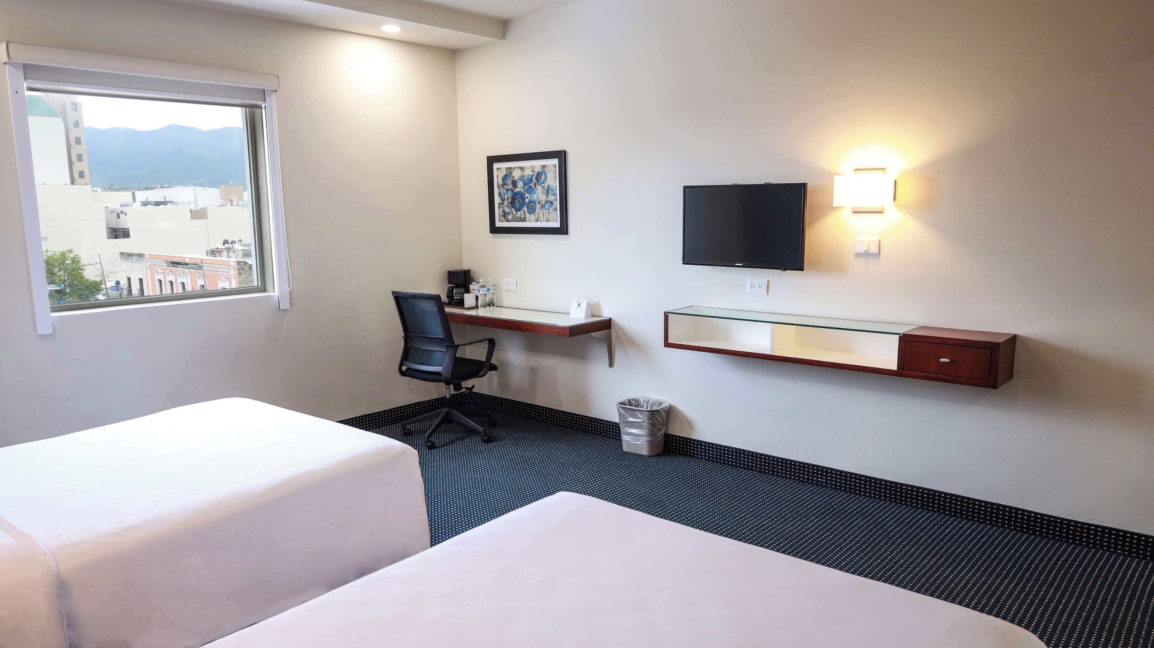 A hotel room with two beds, a desk, a chair, a TV, and a window with a view of buildings and mountains.