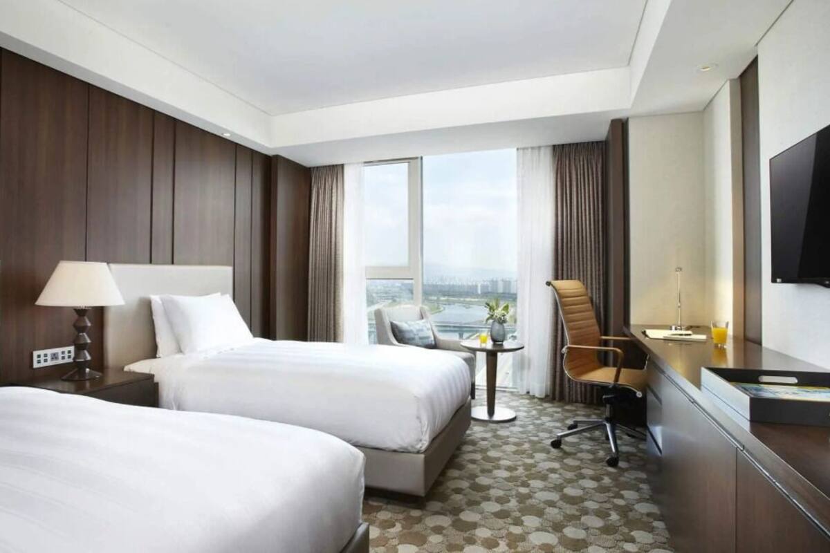 A hotel room with two beds, a desk, a chair, a TV, and a window with a city view.