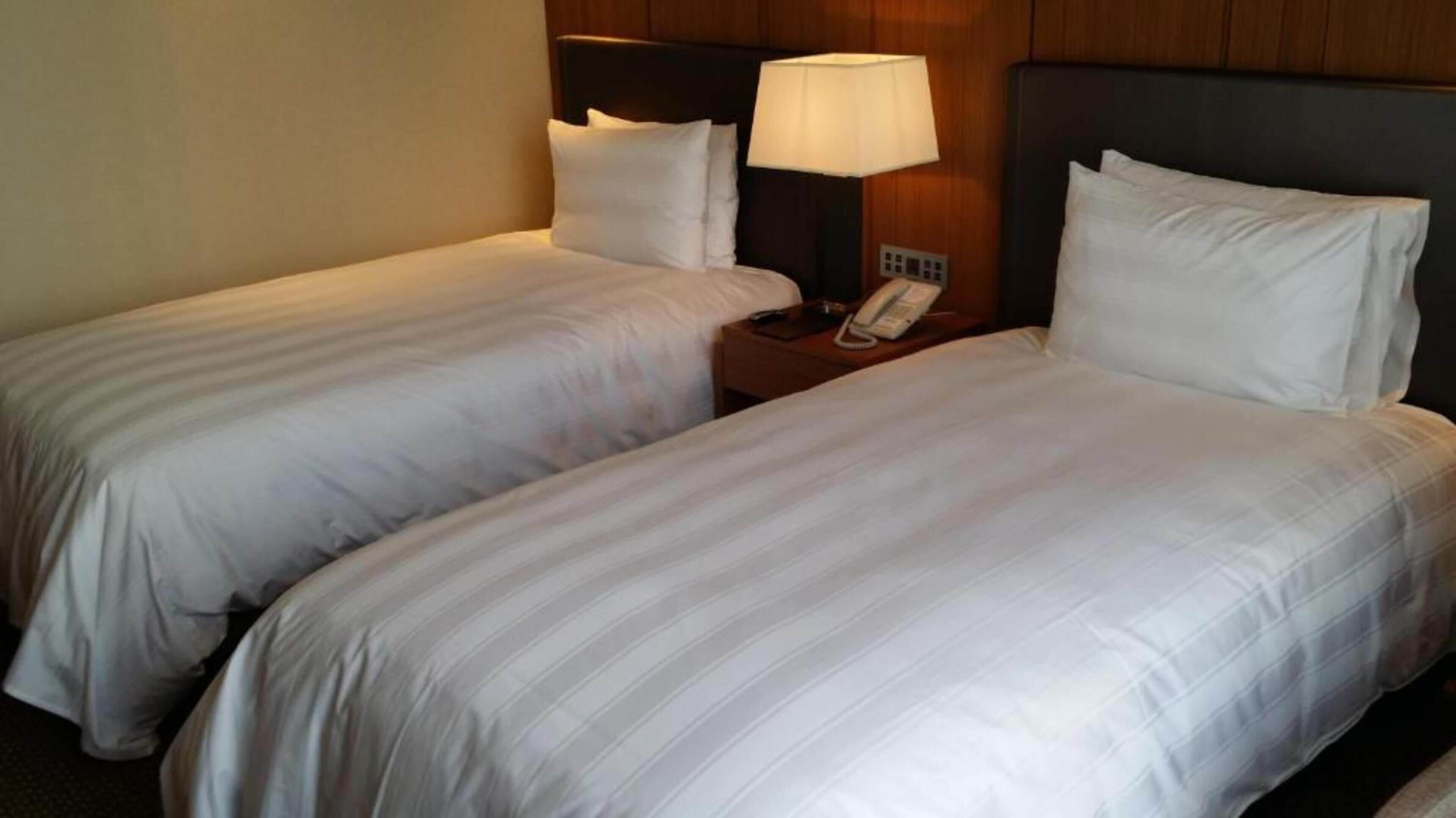 A hotel room with two single beds, each with white linens, a bedside table with a lamp, and a phone.