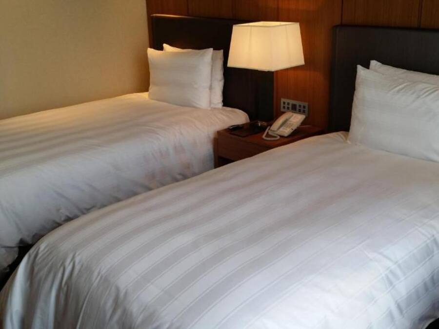 A hotel room with two single beds, each with white linens, a bedside table with a lamp, and a phone.