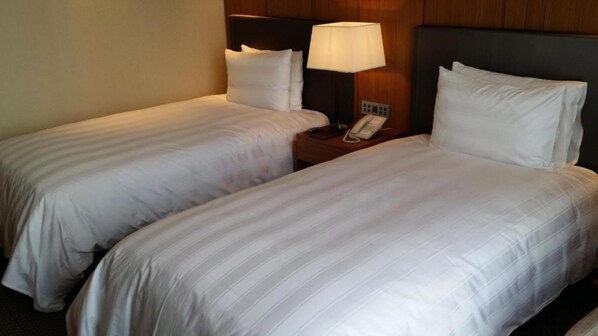 Habitación de hotel con dos camas individuales, cada una con ropa blanca, una mesita de noche con lámpara y teléfono.