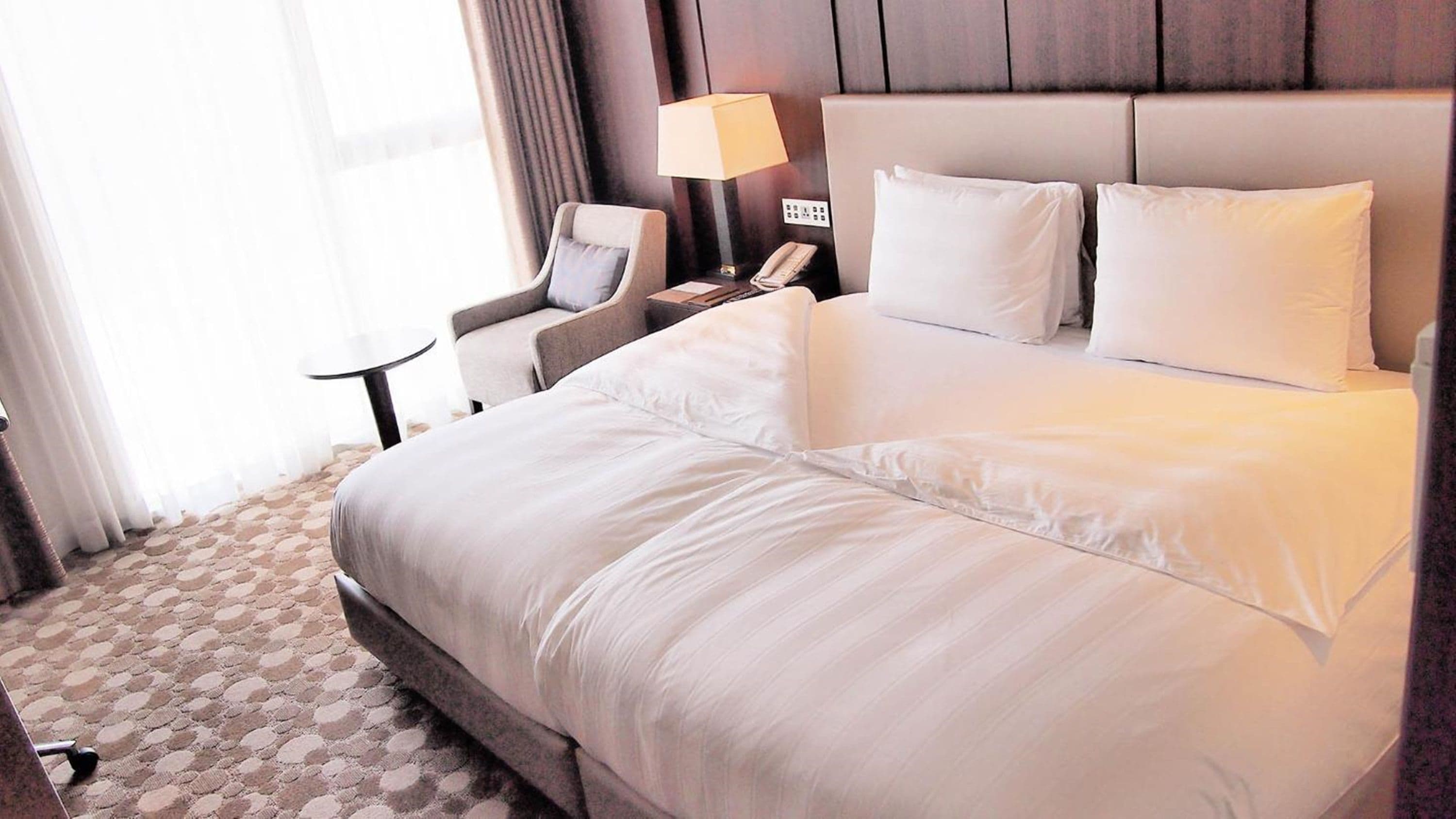 A hotel room with a large bed, a chair, a small table, a lamp, and a phone on the nightstand.