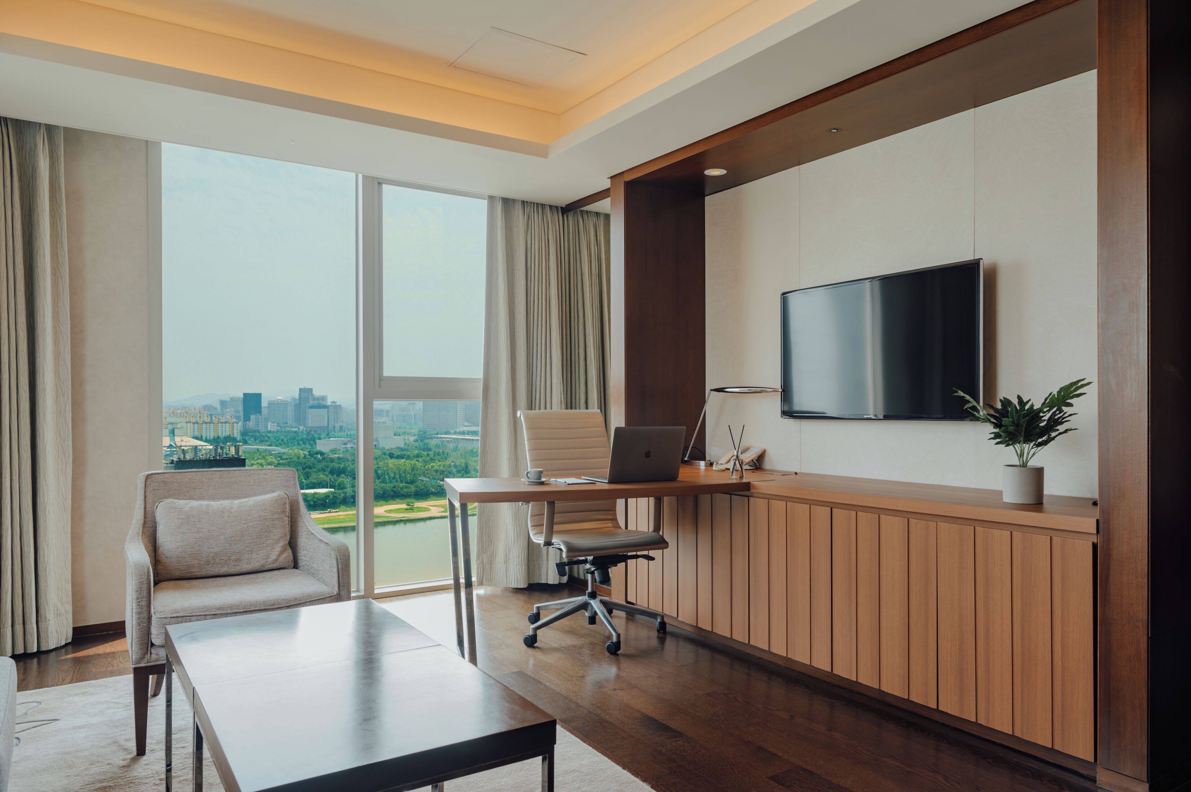 A modern hotel room with a large window, a desk with a laptop, a chair, a TV, and a potted plant.