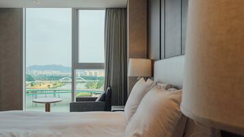 A hotel room with a large bed, a round table, a chair, a lamp, and a view of a cityscape through a large window.
