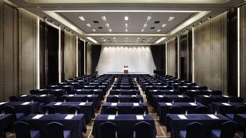 Une salle de conférence avec des rangées de tables et de chaises, un pupitre à l’avant et un rideau en arrière-plan.