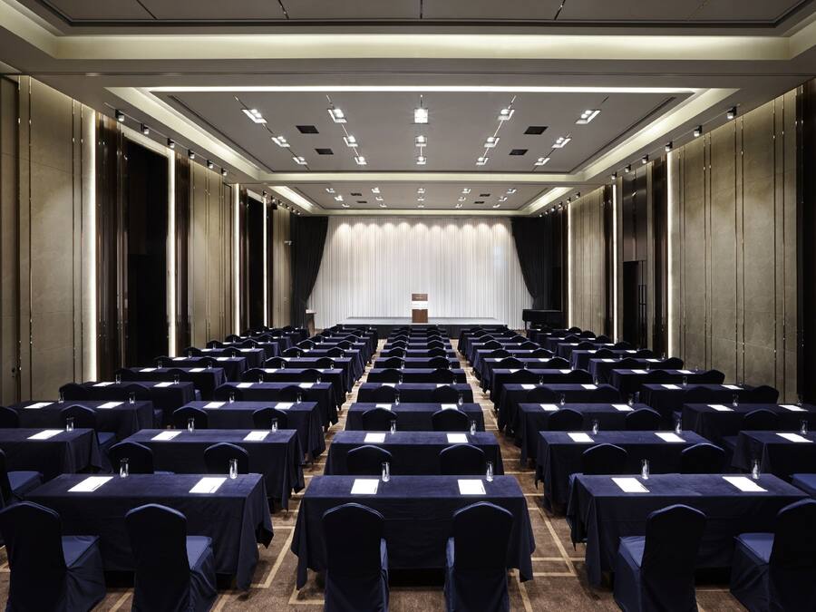 Une salle de conférence avec des rangées de tables et de chaises, un pupitre à l’avant et un rideau en arrière-plan.