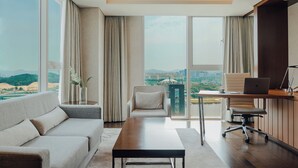 Una sala de estar moderna con un sofá, una mesa de centro, un escritorio con una computadora portátil y vistas a un paisaje urbano a través de ventanas amplias.