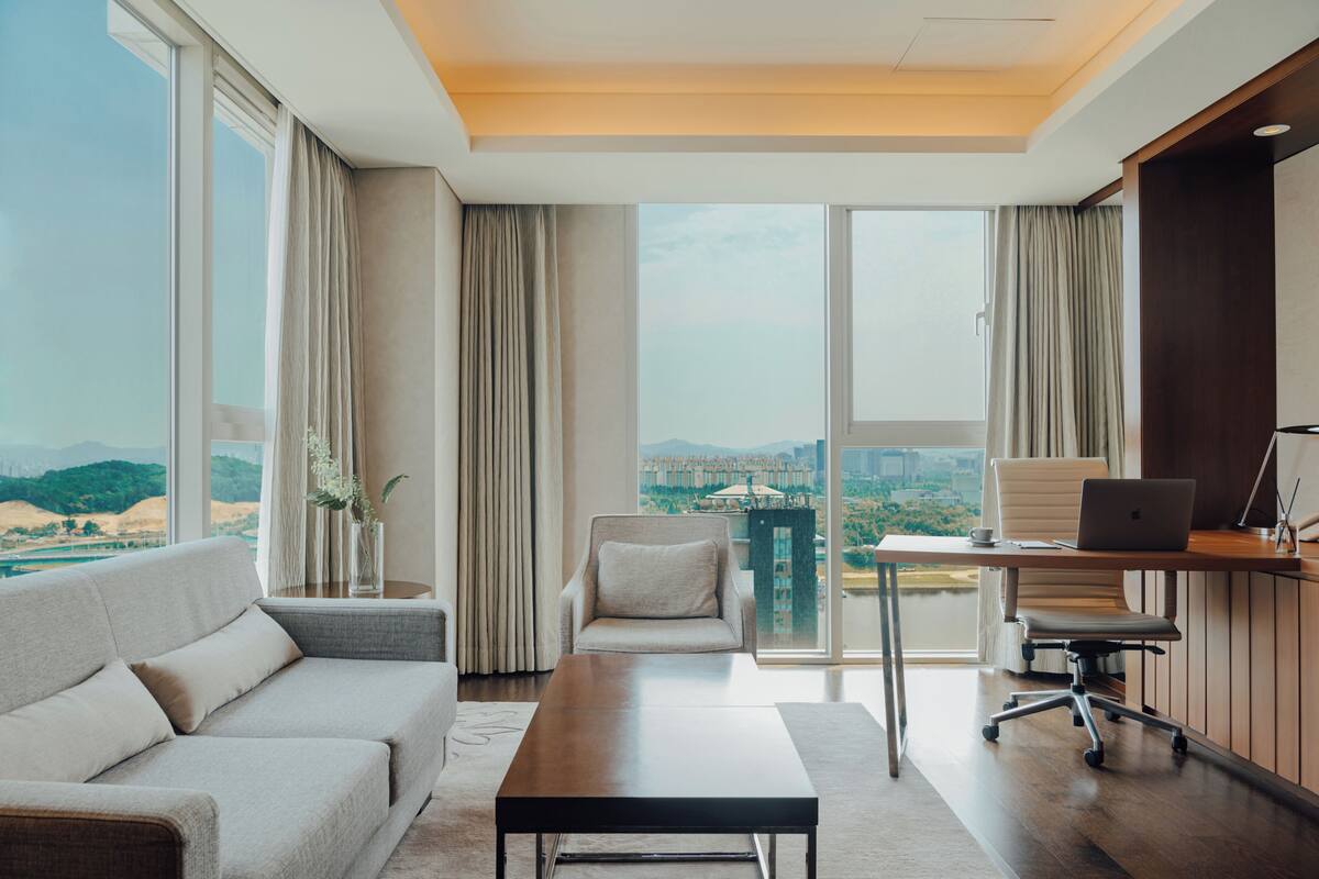 A modern living room with a sofa, a coffee table, a desk with a laptop, and a view of a cityscape through large windows.