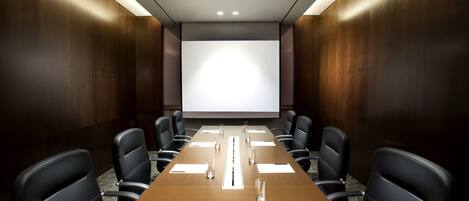 Un salón de conferencias con una larga mesa, sillas negras y una pantalla para proyector.