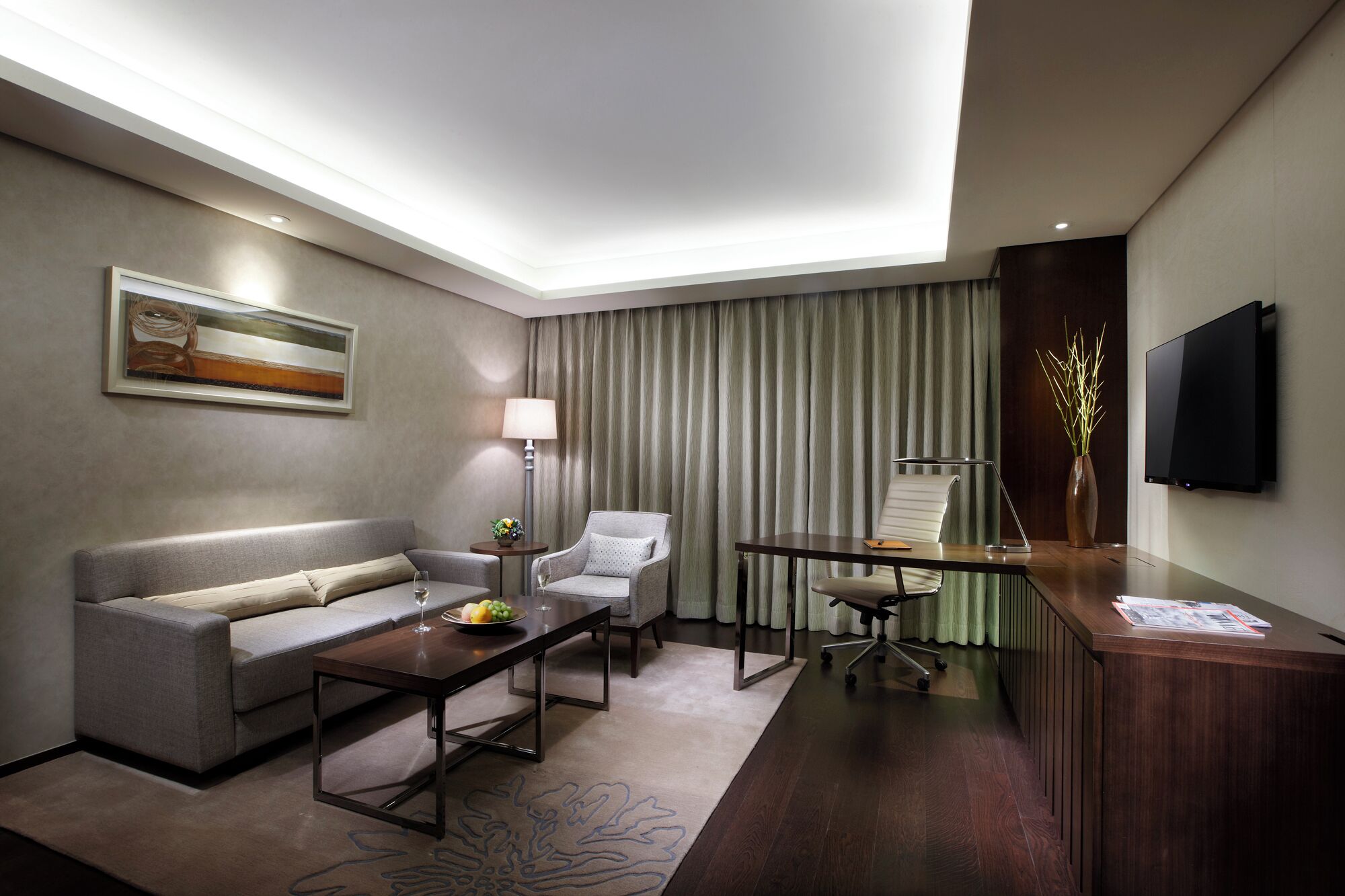 Quarto de hotel moderno com sofá, poltrona, mesa de centro, escrivaninha e televisão de tela plana.