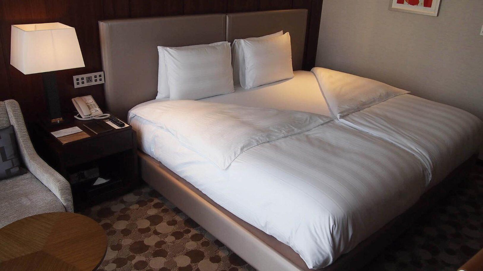 Een net opgemaakt bed met witte lakens, een bedlamp, een telefoon en een kamerbreed tapijt.