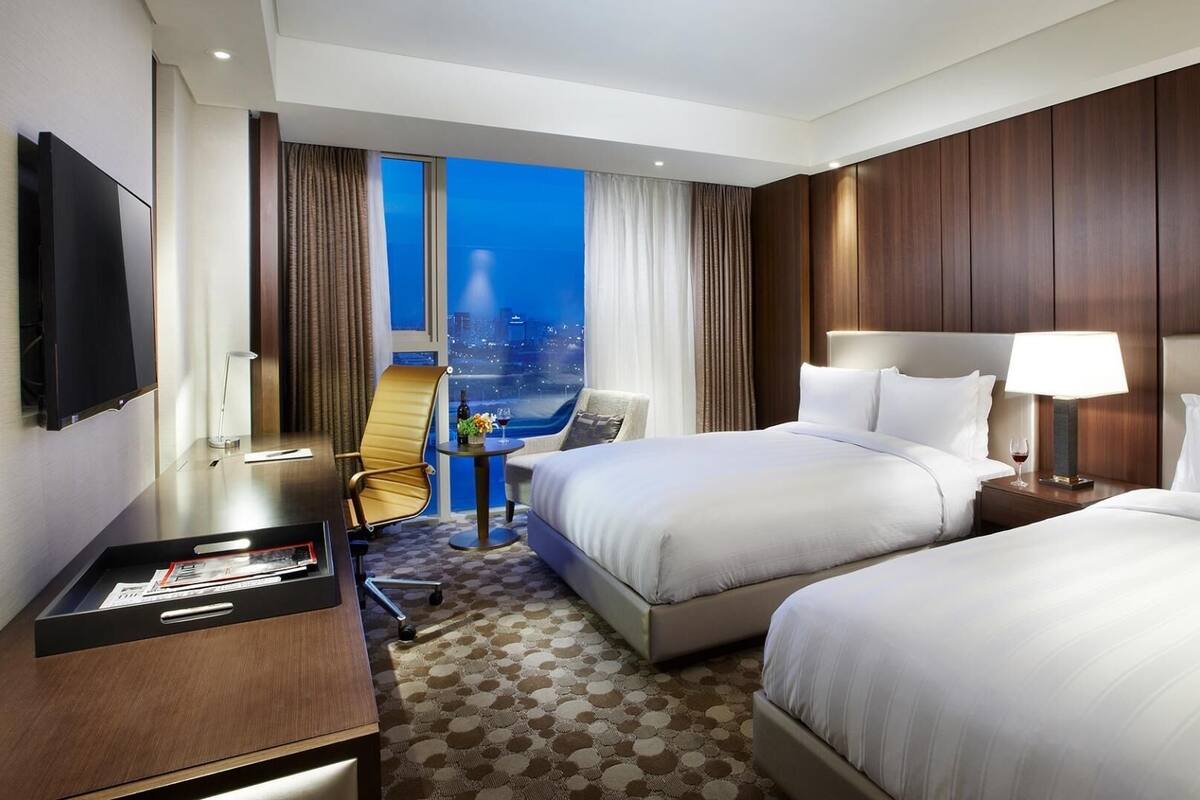 A hotel room with two beds, a desk, a TV, and a view of the city at night.