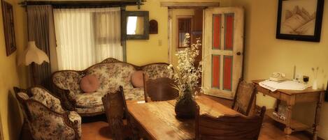 A cozy living room with a floral sofa, wooden chairs, a wooden dining table, and a vase of flowers.