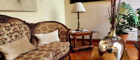 A living room with a floral sofa, a small table with a lamp, and a large vase with plants.