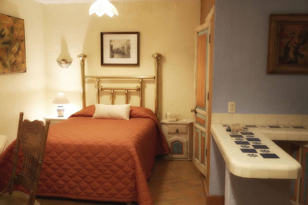 Une pièce comprenant un lit, une chaise, un bureau avec un jeu d’échecs, une lampe et un tableau encadré au mur.