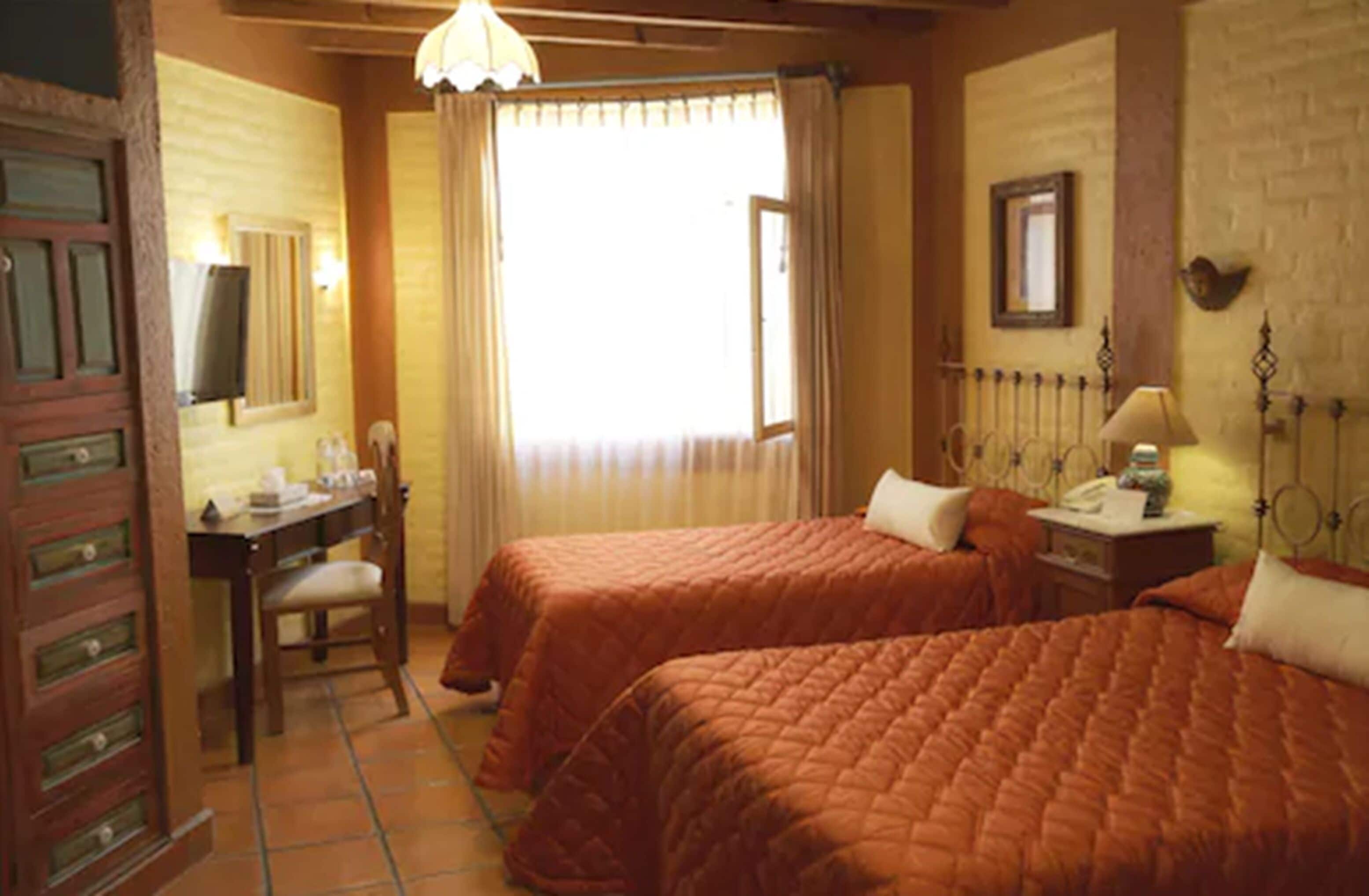 A room with two beds, a desk, a chair, a lamp, and a window with curtains.