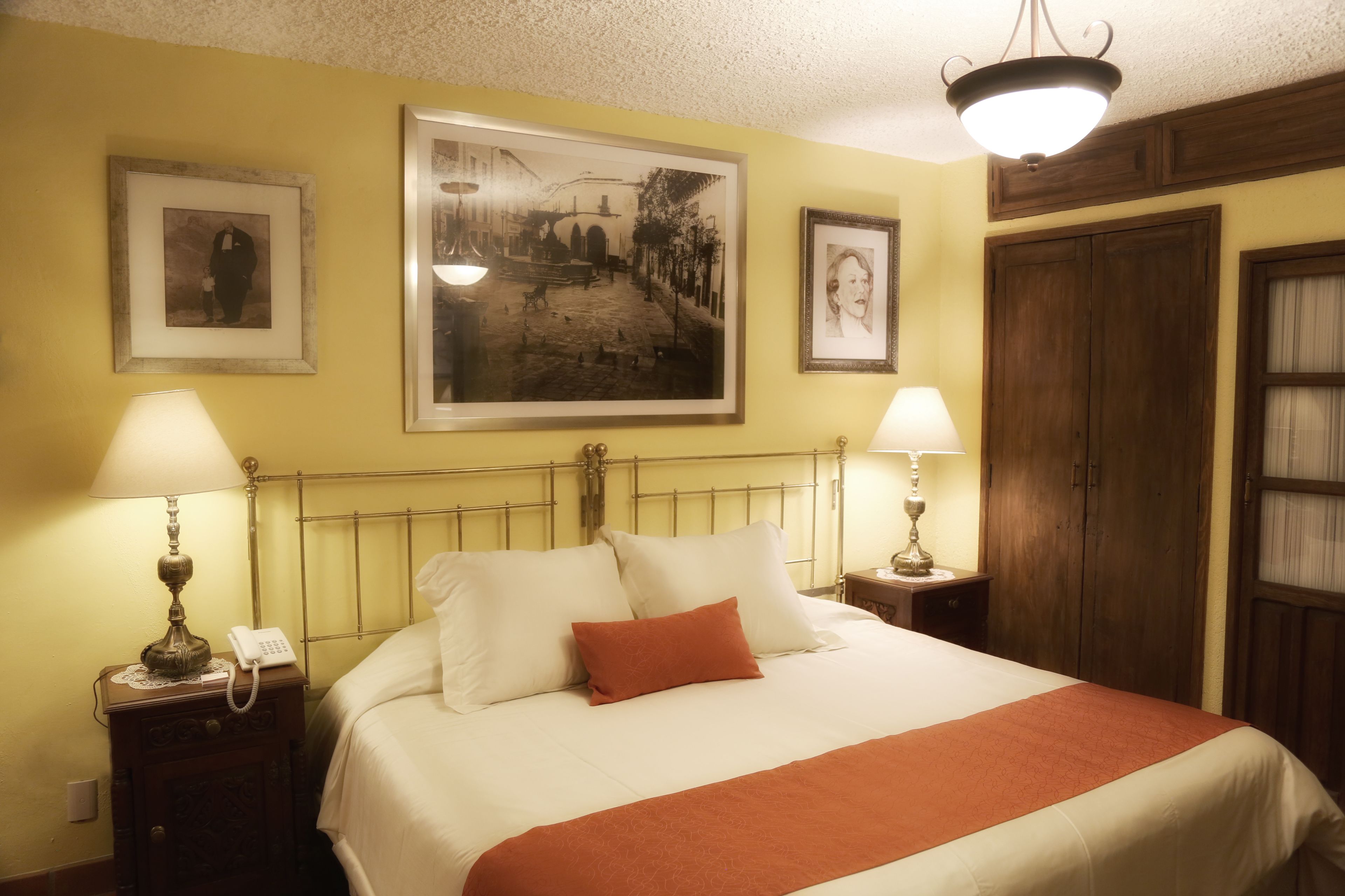 Een net opgemaakt bed met wit en oranje beddengoed, twee bedlampen en lijstjes met foto's aan de muur.