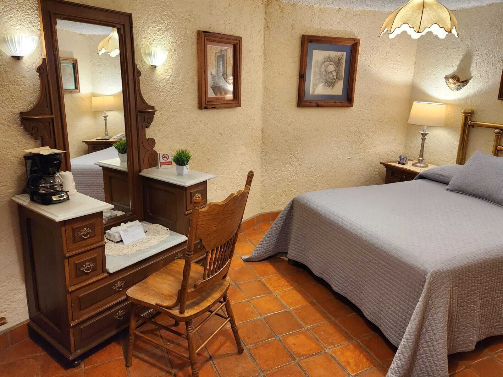 A bedroom with a bed, a wooden chair, a desk with a coffee maker, and two wall-mounted lamps.