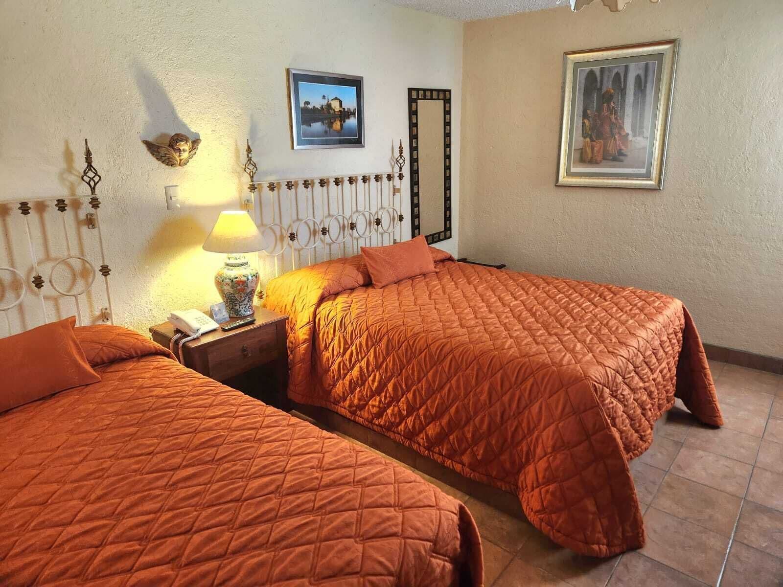 Une chambre d’hôtel avec deux lits, une table de chevet avec une lampe, un miroir et deux tableaux encadrés au mur.