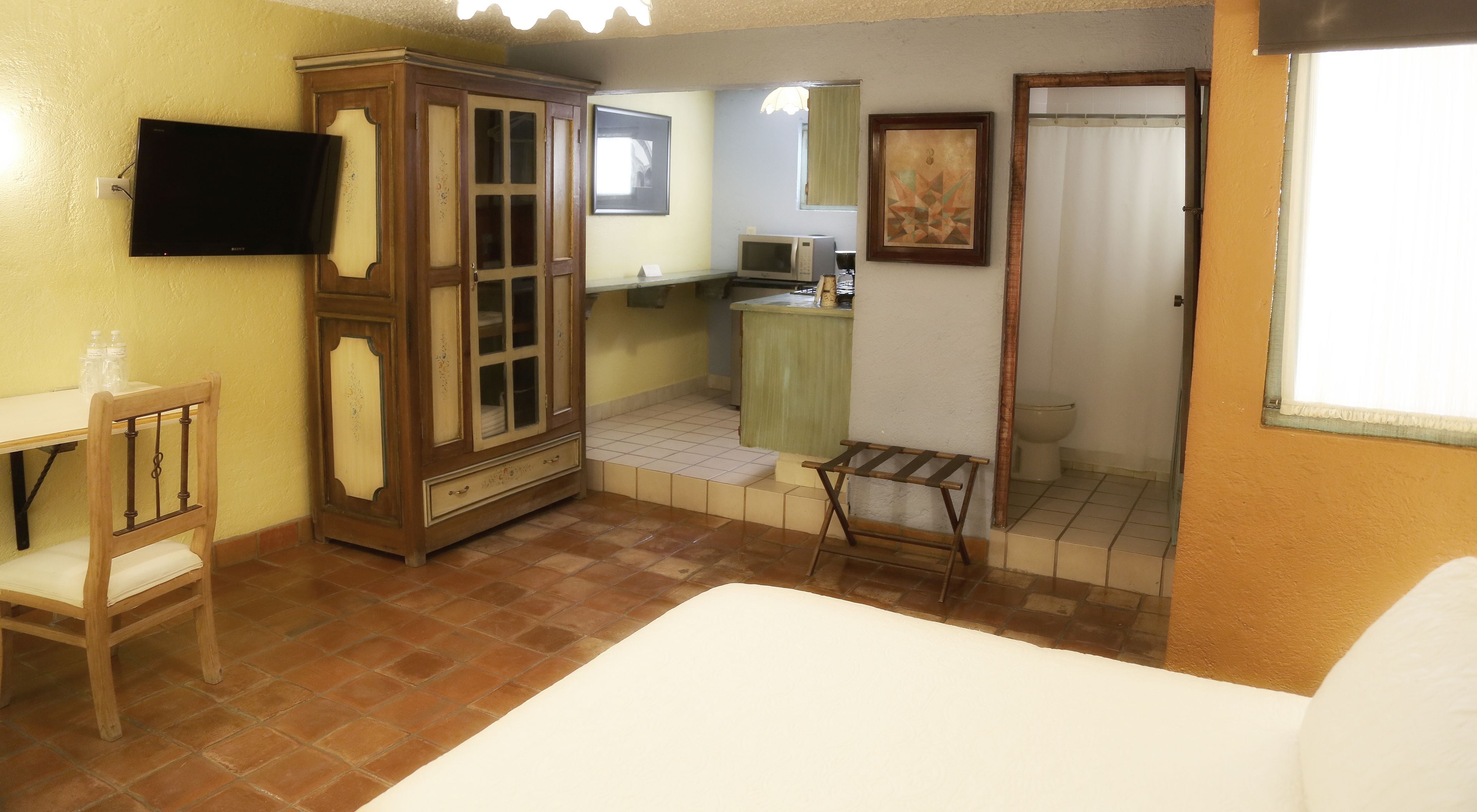 A compact hotel room with a bed, a wooden wardrobe, a small table with chairs, a television, a microwave, a sink, and a toilet.