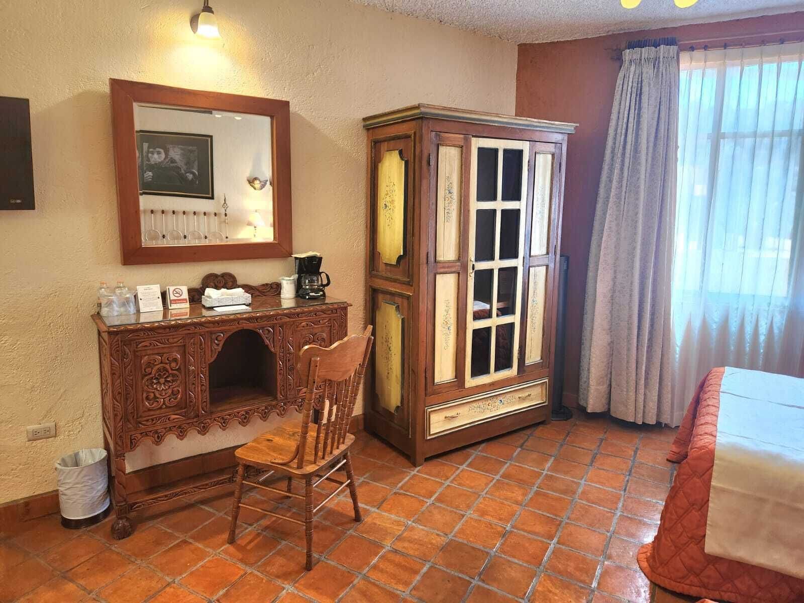 Quarto com escrivaninha de madeira, cadeira e armário, uma cama com cobertor vermelho e uma janela com cortinas.
