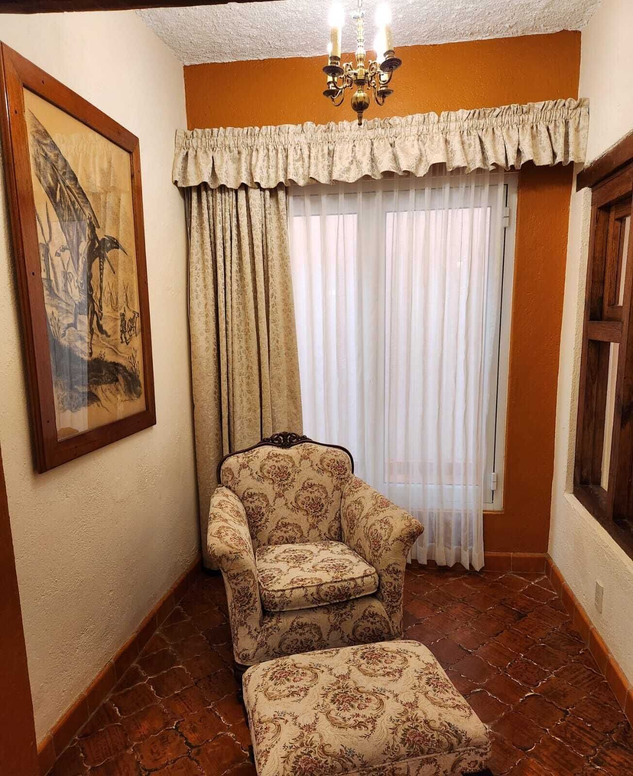 Une pièce d’angle avec un fauteuil et un pouf à motifs, un tableau encadré, un lustre et une fenêtre avec des rideaux.
