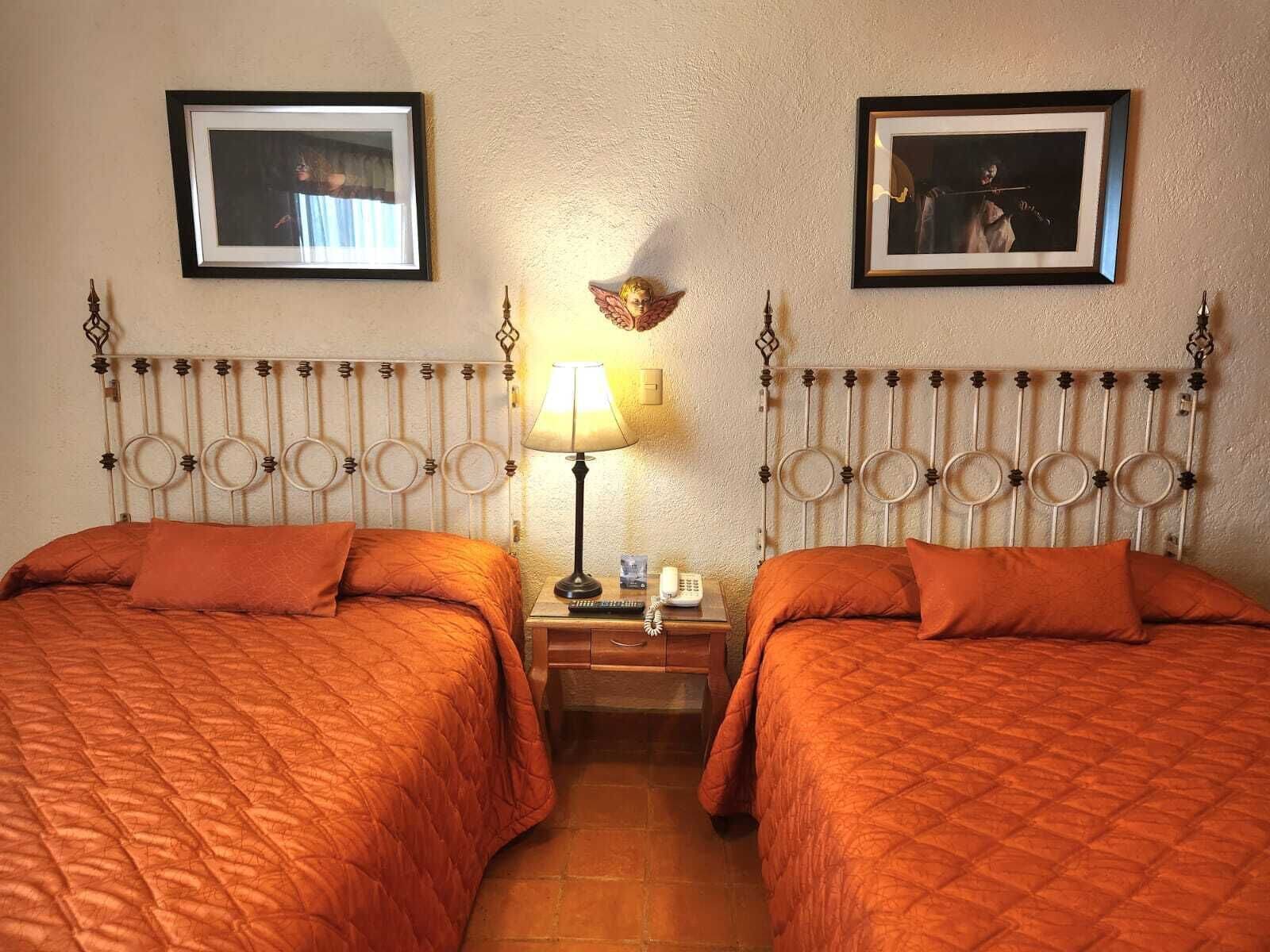 Una habitación con dos camas, cada una con colcha naranja, una mesita de noche con lámpara y cuadros enmarcados en la pared.
