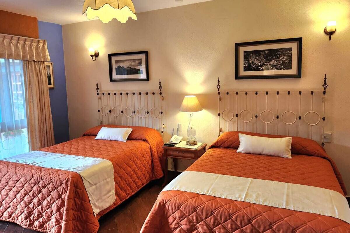Une chambre d’hôtel avec deux lits, chacun recouvert d’une courtepointe orange matelassée et agrémenté d’oreillers blancs. On y trouve une table de chevet avec une lampe, ainsi qu’un tableau encadré accroché au mur.